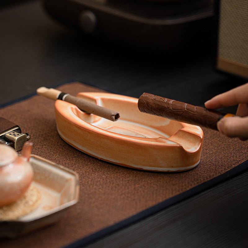 Cigare en terrasse posé sur cendrier céramique Shino terracotta, briquet Zippo en cuir et théière en terre cuite sur tapis tressé naturel, lumière chaude